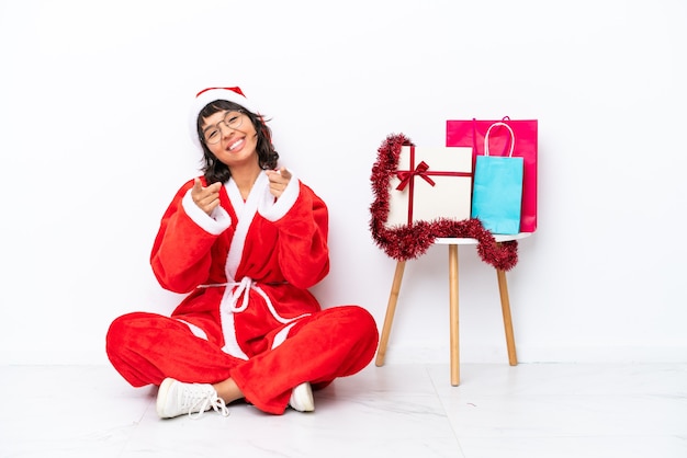 Junges Mädchen, das Weihnachten feiert, das auf dem Boden sitzt, isoliert auf weißem Hintergrund, zeigt mit dem Finger auf dich, während er lächelt