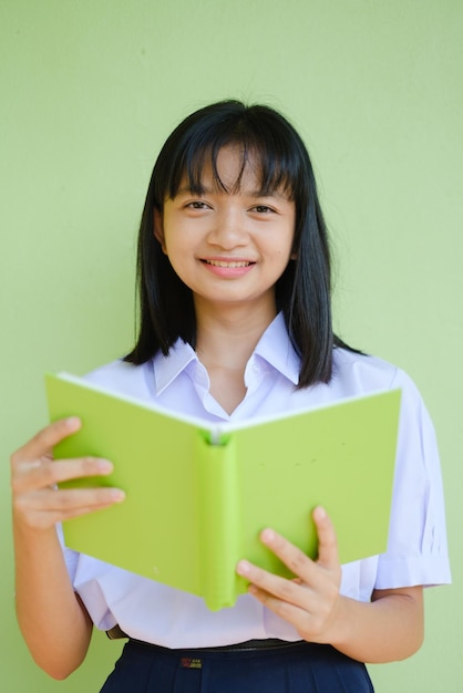 Junges Mädchen des Porträtstudenten in der einheitlichen Schule mit grünem Buch auf grünem Hintergrund, asiatisches Mädchen, Jugendlicher.