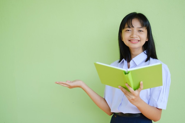 Junges Mädchen des Porträtstudenten in der einheitlichen Schule mit grünem Buch auf grünem Hintergrund, asiatisches Mädchen, Jugendlicher.