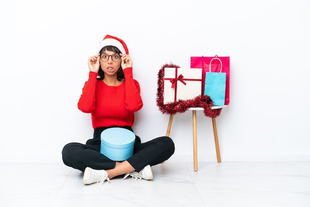 Junges Mädchen feiert Weihnachten auf dem Boden isoliert auf weißem Hintergrund mit Brille und überrascht