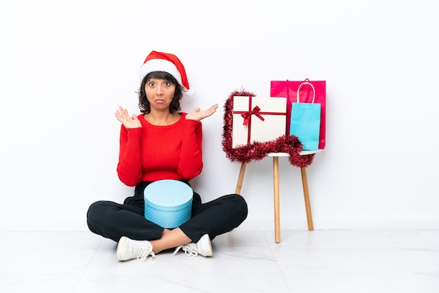 Junges Mädchen feiert Weihnachten auf dem Boden sitzend isoliert auf weißem Hintergrund und hat Zweifel, während sie die Hände hebt