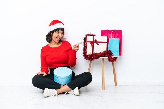 Junges Mädchen feiert Weihnachten auf dem Boden sitzend, isoliert auf weißem Hintergrund, zeigt mit dem Finger zur Seite und präsentiert ein Produkt