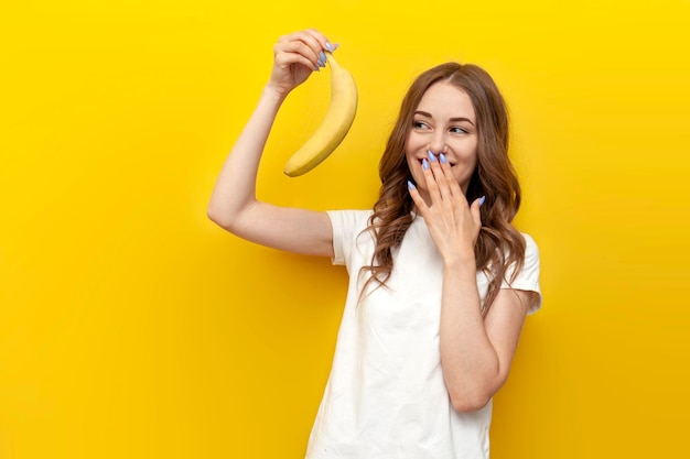 Junges Mädchen hält eine kleine Banane in der Hand und macht sich auf gelbem, isoliertem Hintergrund lustig. Frau präsentiert Erotik