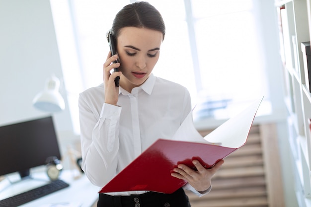 Junges Mädchen im Büro nahe dem Gestell und Blättern durch den Ordner mit den Dokumenten und spricht am Telefon.