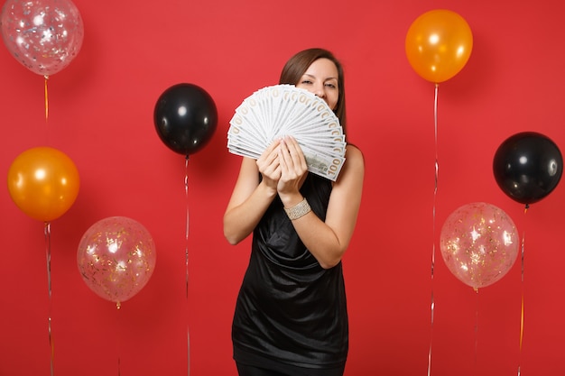 Junges Mädchen im schwarzen Kleid versteckt, Gesicht mit Bündel viel Dollar, Bargeld in der Hand auf leuchtend roten Hintergrundluftballons bedeckend. Frauentag Happy New Year Geburtstag Mockup Urlaub Party Konzept.