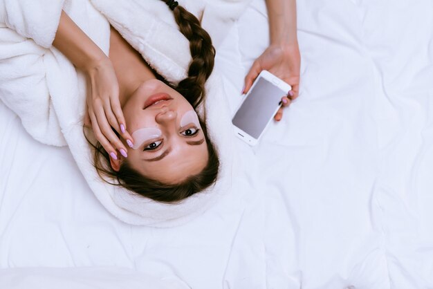 Junges Mädchen im weißen Gewand liegt im Bett, unter Augenflecken, hält Telefon