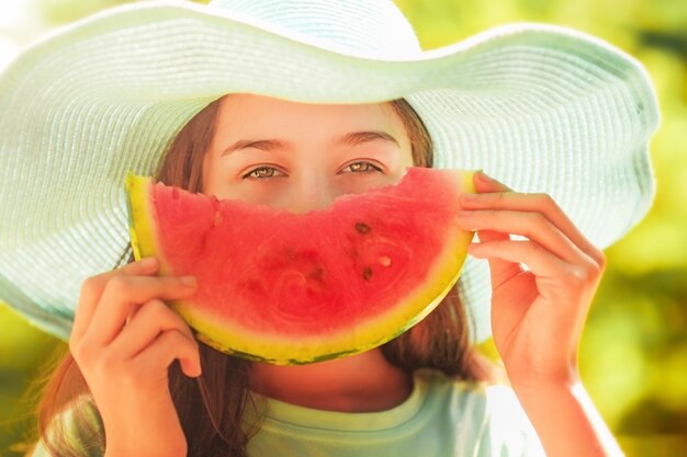 Junges Mädchen in einem blauen Hut, der eine Scheibe Wassermelone auf einem Sommerhintergrund hält. Vegetarier