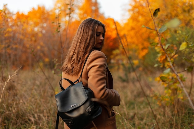 Junges Mädchen in einem Herbstmantel mit einem Lederrucksack reist auf einer Natur im Wald mit gelbem Herbstlaub