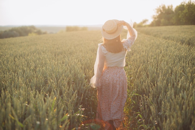 Junges Mädchen in einem schönen langen Kleid und Hut geht mit Weizen in der Hand auf einem Feld spazieren