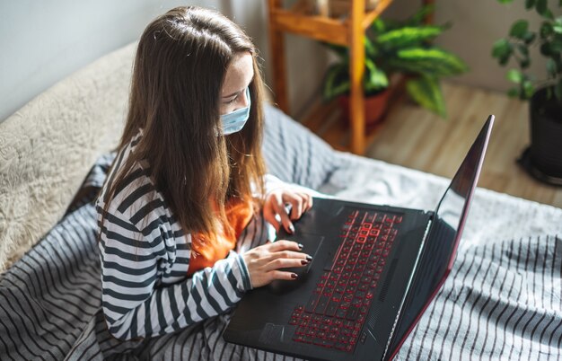 Junges Mädchen in einer Schutzmaske sitzt auf einem Bett zu Hause und benutzt einen Laptop
