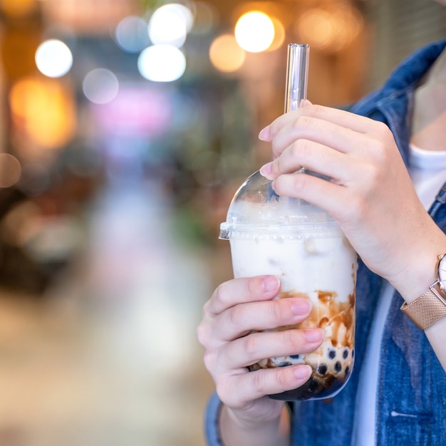 Junges Mädchen in Jeansjacke trinkt Tapioka-Perlen-Blasenmilchtee mit braunem Zuckergeschmack mit Glasstroh auf dem Nachtmarkt von Taiwan, Nahaufnahme, Bokeh