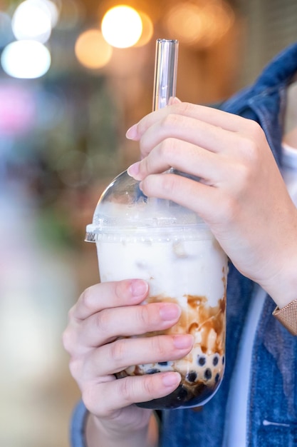 Junges Mädchen in Jeansjacke trinkt Tapioka-Perlenblasenmilchtee mit braunem Zuckergeschmack und Glasstroh auf dem Nachtmarkt von Taiwan, Nahaufnahme Bokeh