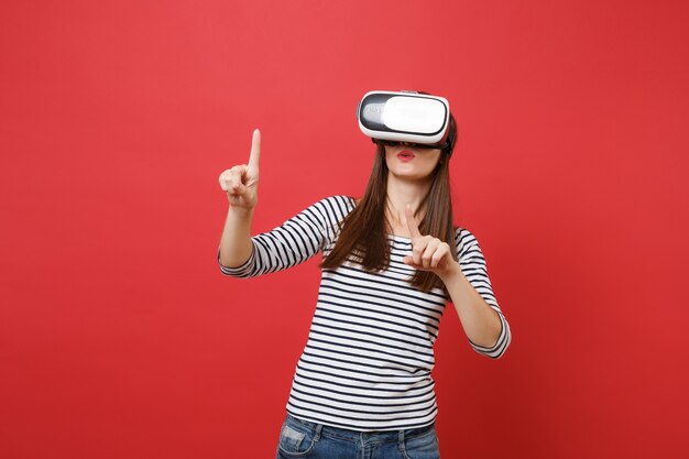 Junges Mädchen in Virtual-Reality-Brille berührt so etwas wie den Knopfdruck, der auf den schwebenden virtuellen Bildschirm zeigt, der auf rotem Hintergrund isoliert ist. Menschen aufrichtige Emotionen Lifestyle-Konzept. Kopieren Sie Platz.