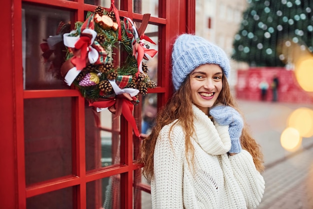 Junges Mädchen in warmer Kleidung macht einen Spaziergang im Freien in der Stadt in der Nähe der roten Telefonstation.