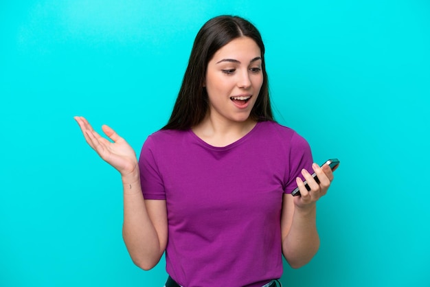 Junges Mädchen isoliert auf blauem Hintergrund, das die Kamera anschaut, während es das Handy mit überraschtem Ausdruck benutzt