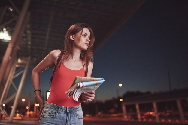 Junges Mädchen kostet nachts in der Nähe des Terminals des Flughafens oder Bahnhofs und liest Stadtplan