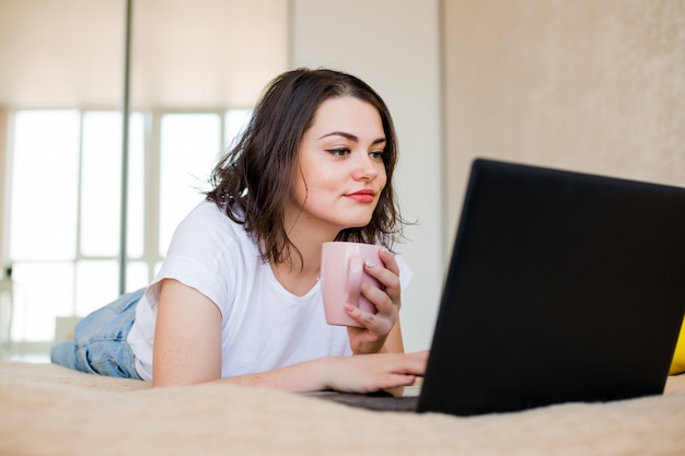 Junges Mädchen liegt zu Hause auf einem Bett mit Kaffee und einem Laptop