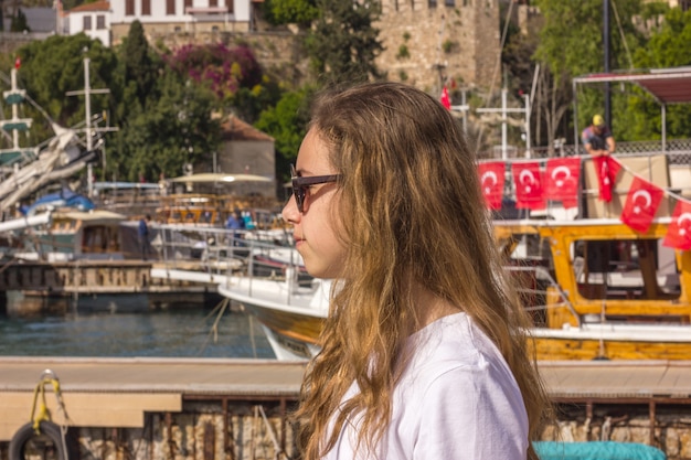 Junges Mädchen mit dem langen Haar sitzt auf dem Pier im Yachthafen und betrachtet das blaue Meer