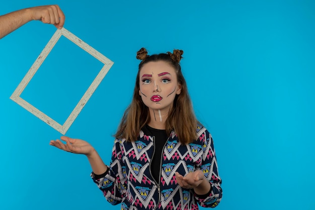 Junges Mädchen mit Fantasie-Make-up, das leeren Rahmen auf blauem Hintergrund hält. Foto in hoher Qualität