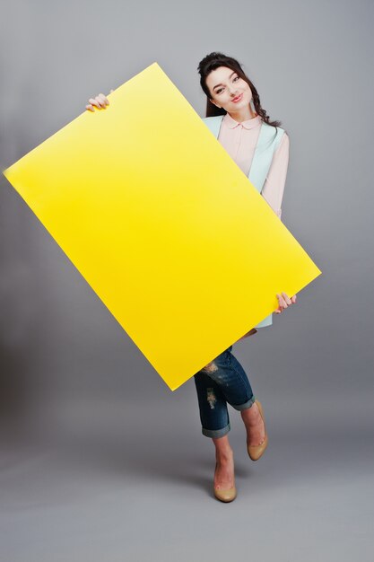 Junges Mädchen mit gelbem leerem Papier des Gesichtsgriffs. Leere Karte der Show der jungen Frau. Mädchen mit dem langen Haarporträt lokalisiert auf grauem Hintergrund.