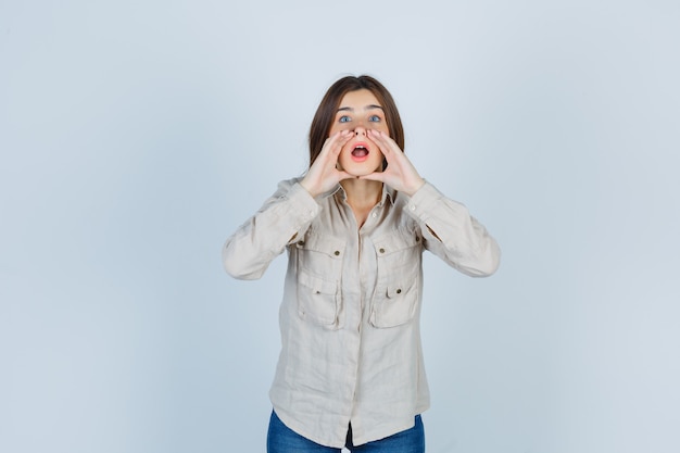 Junges Mädchen mit Händen in der Nähe des Mundes, als jemanden in beigefarbenem Hemd, Jeans anzurufen und fokussiert zu schauen, Vorderansicht.