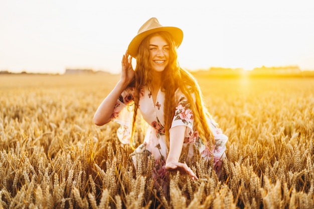 Junges Mädchen mit langem lockigem Haar und Sommersprossengesicht, im Hut, im hellen weißen Kleid mit Blumendruck, stehend im Weizenfeld, posierend für Kamera, im Hintergrund Sonnenuntergang.