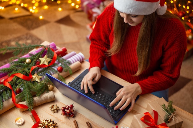 Junges Mädchen mit Laptop sitzt am Tisch mit vielen Geschenken und Dekorationen Junges Mädchen bestellt Weihnachtsgeschenke für Freunde und Verwandte, die im Internet von Online-Shops einkaufen