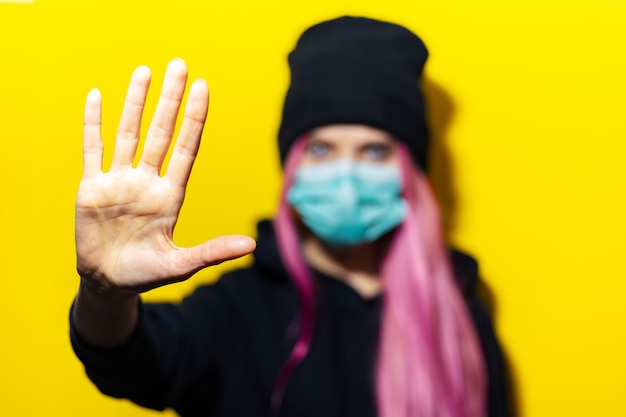 Foto junges mädchen mit rosa haaren und blauen augen, die medizinische grippemaske tragen, gekleidet in schwarzen kapuzenpullover und mützenhut auf gelber wand.