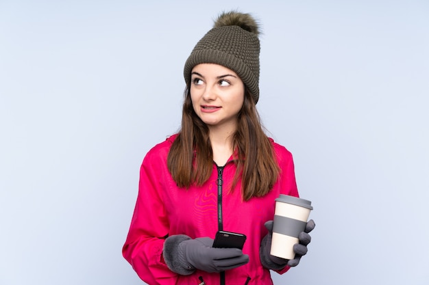 Junges Mädchen mit Wintermütze auf blauer Wand, die Kaffee hält, um und ein Handy wegzunehmen, während sie etwas denken