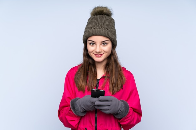 Junges Mädchen mit Wintermütze lokalisiert auf Blau, das eine Nachricht mit dem Handy sendet