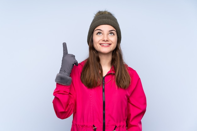 Junges Mädchen mit Wintermütze lokalisiert auf blau nach oben zeigend und überrascht