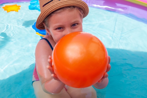 Junges Mädchen spielt im aufblasbaren Wasserschwimmbecken mit buntem Regenbogen und orangefarbenem Ball. Fröhliches Kind mit Strohhut lächelt