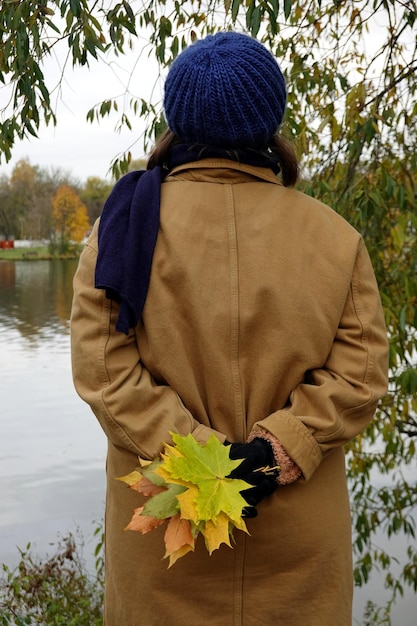 Junges Mädchen trägt im Herbst einen Baumwollmantel und hält hinter dem Rücken einen Bouquet aus bunten Ahornblättern