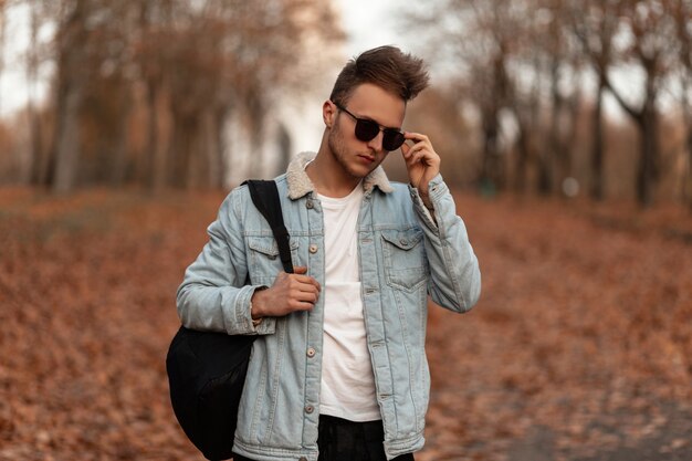 junges Mannmodell in einer stilvollen blauen Jeansjacke, die im Park aufwirft