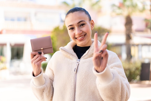 Junges marokkanisches Mädchen, das draußen eine Brieftasche hält, lächelt und zeigt Victory-Zeichen