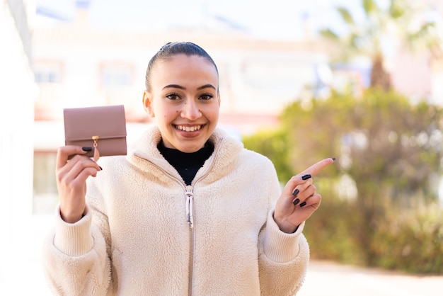 Junges marokkanisches Mädchen, das draußen eine Brieftasche hält, überrascht und zeigt mit dem Finger zur Seite