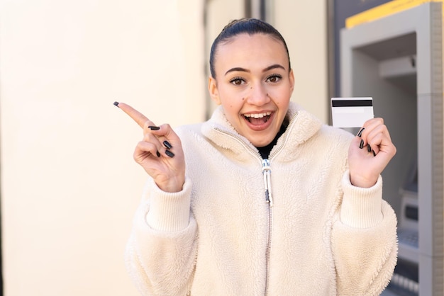 Junges marokkanisches mädchen, das draußen eine kreditkarte hält, überrascht und zeigt mit dem finger zur seite