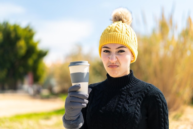 Foto junges marokkanisches mädchen, das wintermuffs trägt, während es draußen einen kaffee mit traurigem ausdruck hält