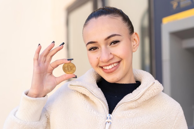 Junges marokkanisches Mädchen im Freien mit einem Bitcoin mit fröhlichem Gesichtsausdruck