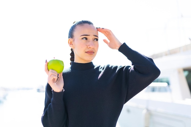 Junges marokkanisches Mädchen mit einem Apfel im Freien, das Zweifel hat und einen verwirrten Gesichtsausdruck hat