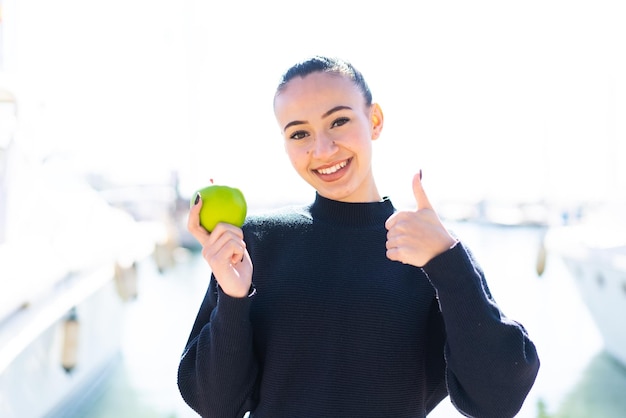 Junges marokkanisches Mädchen mit einem Apfel im Freien mit Daumen nach oben, weil etwas Gutes passiert ist