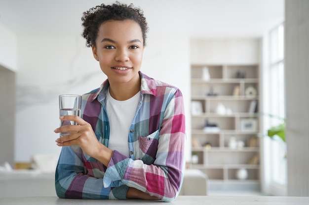 Junges Mischlingsmädchen, das zu Hause ein Glas reines Wasser hält Gesunder Lebensstil, Selbstpflege