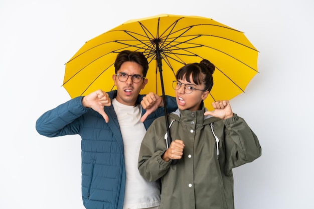 Junges Mischlingspaar, das einen Regenschirm isoliert auf weißem Hintergrund hält und mit beiden Händen den Daumen nach unten zeigt