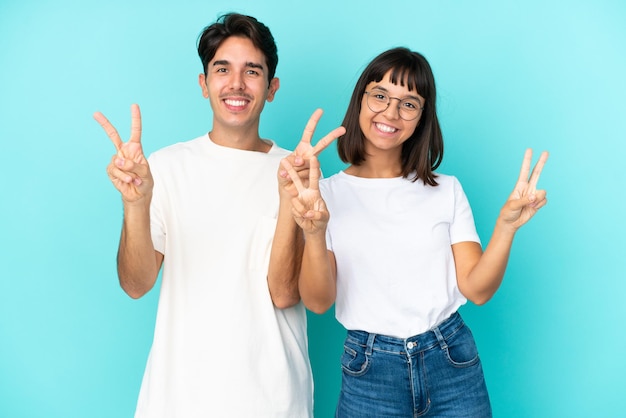 Junges Mischlingspaar isoliert auf blauem Hintergrund, lächelnd und mit beiden Händen Victory-Zeichen zeigend