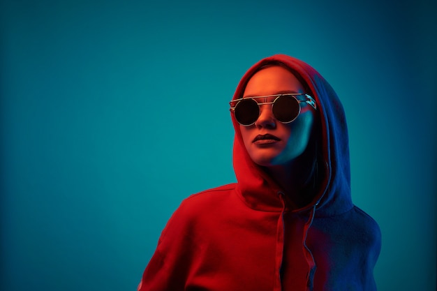 Junges Modell, das in einem Studio in einem trendigen Neonlicht aufwirft. Frau in der runden Sonnenbrille und im Kapuzenpulli mit bunten Lichteffekten.