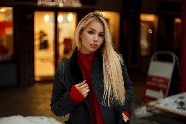 Junges Modell der blonden Frau im Mode-Weinlesemantel mit rotem Pullover mit Tasche, die in der Stadt am Abend geht