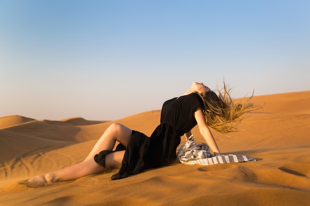 Junges Modell im schwarzen Kleid, das auf dem Sand in der Wüste liegend aufwirft