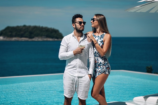 Junges Modepaar mit Sonnenbrille, das an einem sonnigen Sommertag in der Nähe des Infinity-Pools steht. Reise zum warmen Ziel. Flitterwochen. Schöne Meereslandschaft. Phuket. Thailand.