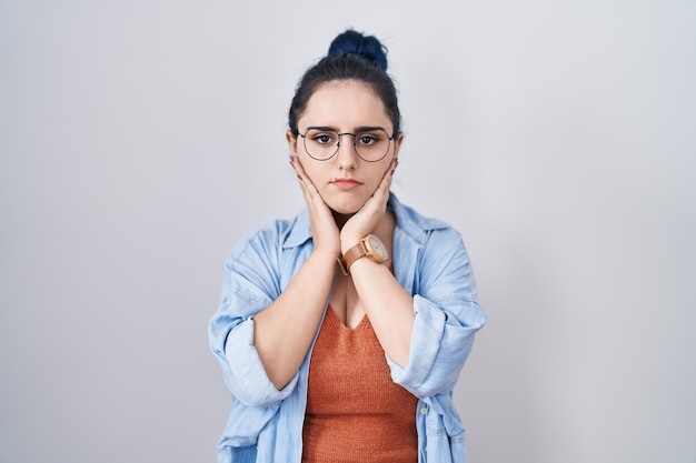 Junges, modernes Mädchen mit blauen Haaren, das vor weißem Hintergrund steht, müde Hände, die das Gesicht bedecken, Depressionen und Traurigkeit, verärgert und gereizt über das Problem