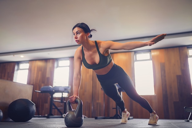 Junges motiviertes Mädchen, das Plankenübung unter Verwendung von Kettlebells mit einer Hand im Fitnessstudio, Foto in voller Länge, Kopierraum tut